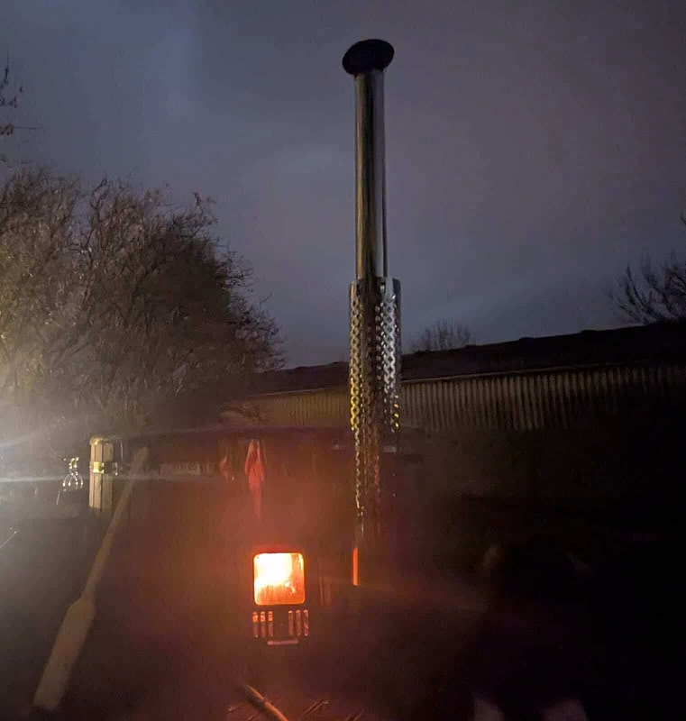 Scène nocturne extérieure montrant la cheminée incandescente d'un poêle à bois avec un écran thermique métallique à côté d'arbres dénudés et de la silhouette sombre d'un bâtiment
