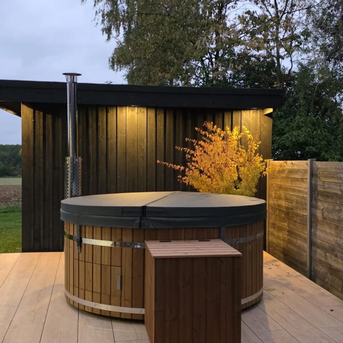 Vue extérieure en soirée d'un bain nordique rond en bois fermé, avec abri près d'un hangar noir et d'un arbre illuminé