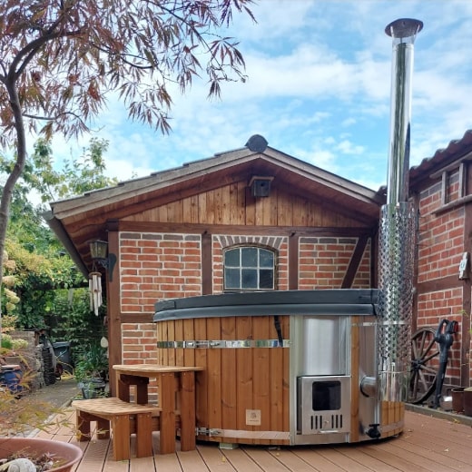 Vue extérieure d'un bain nord-américain rond chauffé au bois, avec poêle en acier inoxydable et marches en bois sur une terrasse en bois près d'un bâtiment en briques