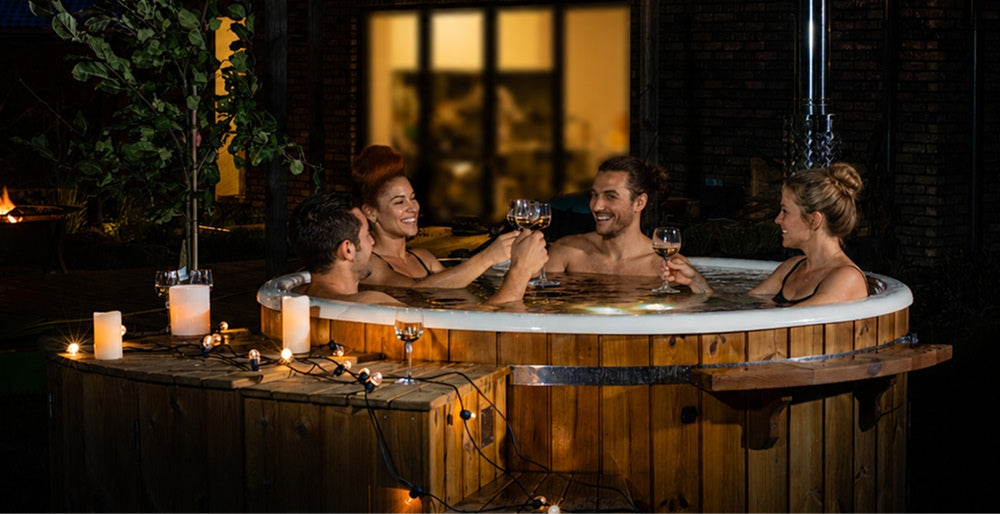 Four friends are soaking in a hot tub on a dark evening and raising their glasses under romantic lighting