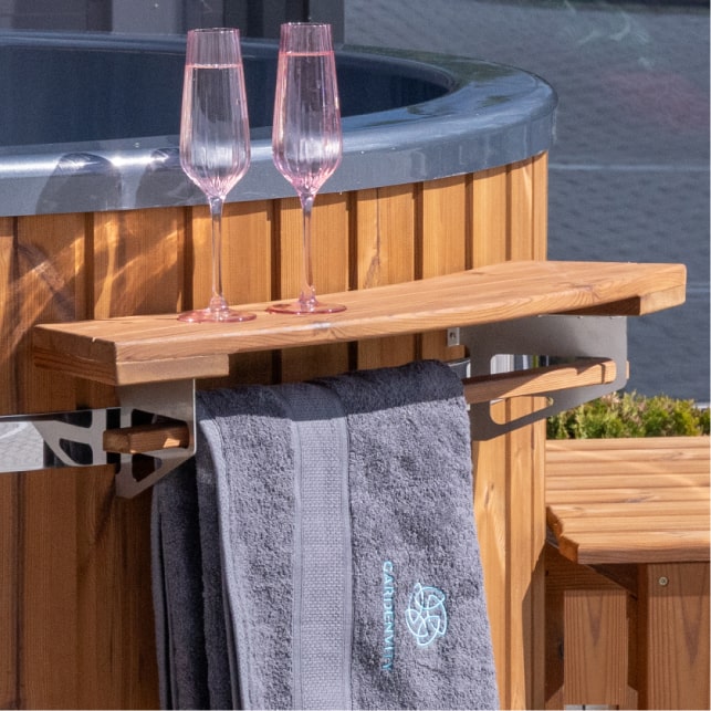 Gros plan sur une étagère en bois fixée à bain nordique, avec deux flûtes à champagne rosé et une serviette grise suspendue en dessous.
