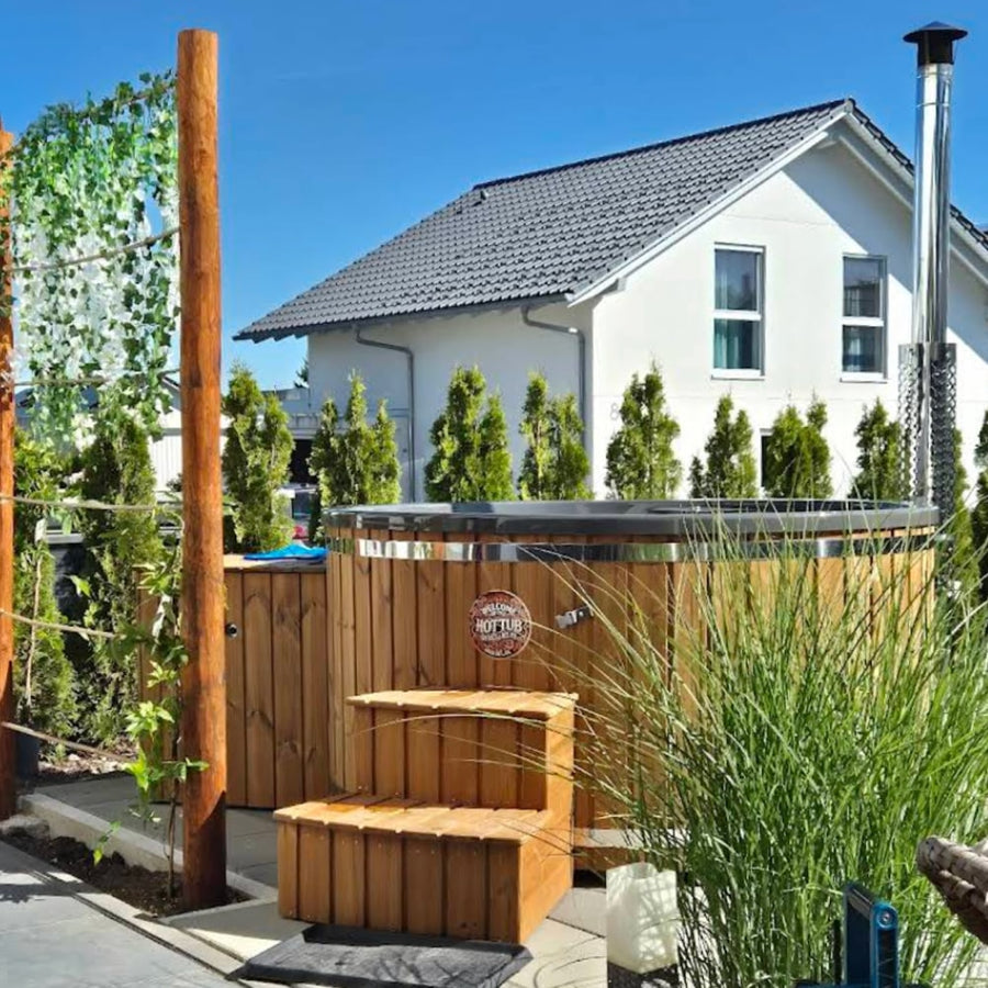 Vue extérieure d'un bain nord-américain chauffé au bois, avec bordure métallique, marches en bois et hautes plantes de confidentialité, à côté d'une maison blanche moderne.