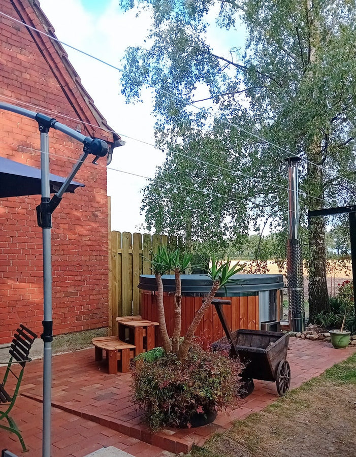 Outdoor view of a wood-fired hot tub with wooden steps, metal chimney, potted plants, and brick patio next to a red brick building