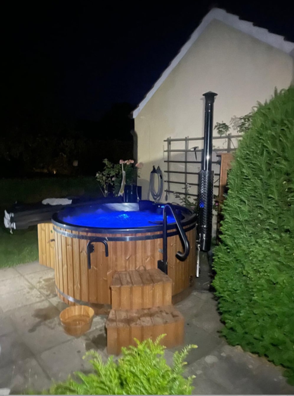 Night view of wooden hot tub with blue illuminated water, wooden steps, stainless steel stove pipe, and garden surroundings