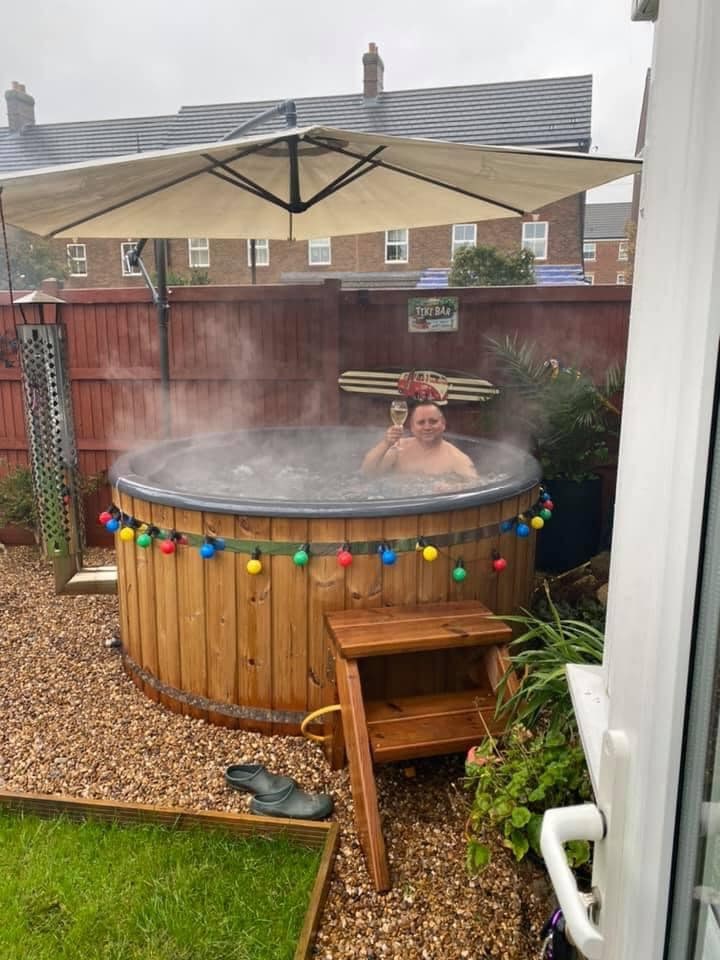 Outdoor view of a round wood-fired hot tub with wooden steps, colorful lights, large umbrella, and steam rising in backyard setting
