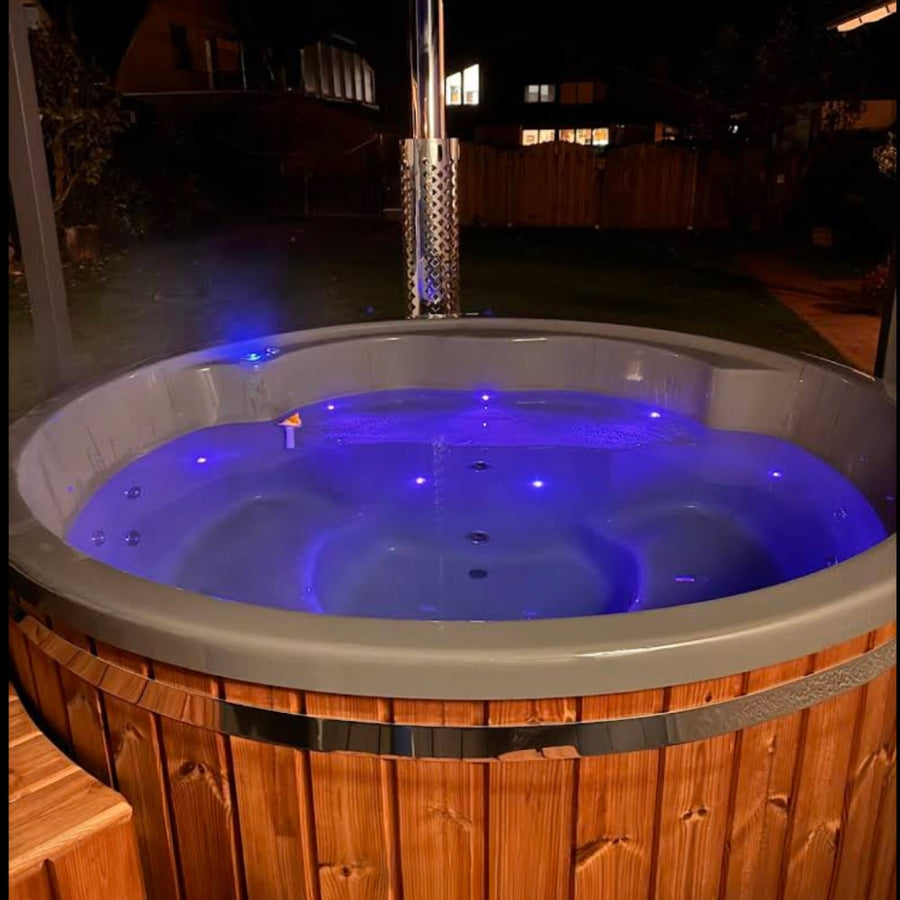 Vue nocturne d'un bain nord-nordique illuminé au feu de bois avec des lumières LED bleues et une cheminée en acier inoxydable dans un cadre extérieur