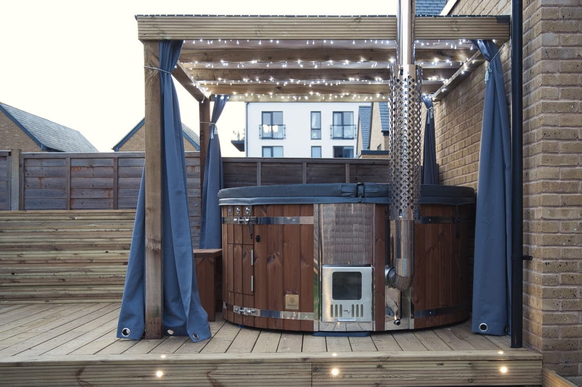 Gardenvity wood fired hot tub on wooden decking under pergola with string lights in a modern courtyard.