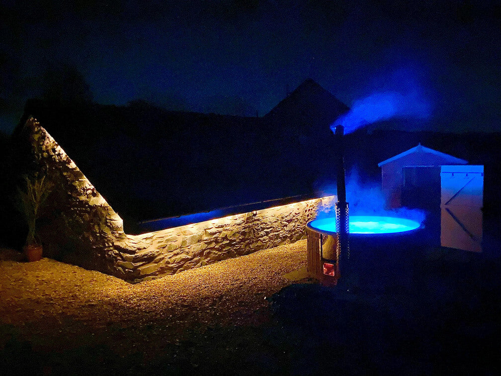Nighttime outdoor scene with a glowing blue illuminated hot tub beside a stone wall and shed in darkness