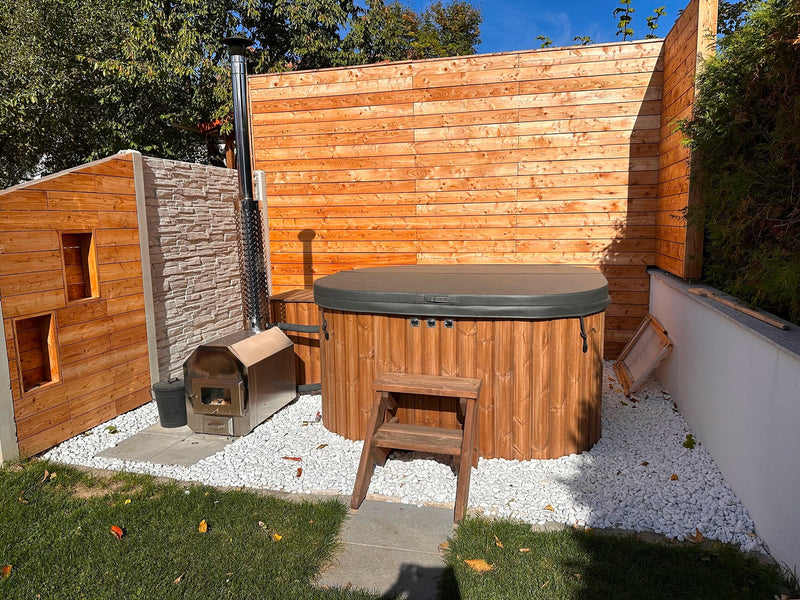 Outdoor corner view of wood fired hot tub with cover, small wooden steps, metal stove, and wood panel privacy walls