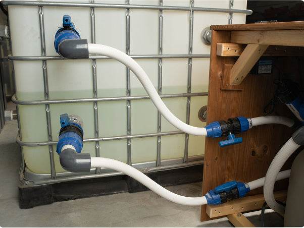 Close-up view of flexible white pipes with blue valves connected between a large plastic water tank and wooden structure.