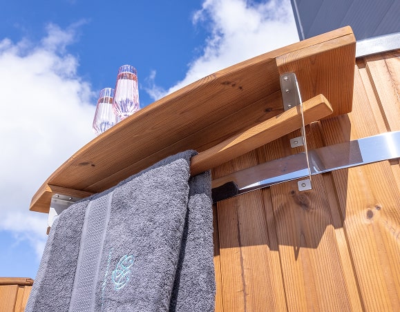 Gros plan sur un porte-serviettes extérieur en bois fixé à un bain-marie chauffé au bois, contenant deux serviettes grises sous un ciel bleu.