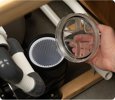 Close-up of hand removing clear lid to reveal clean filter inside plumbing system under wooden sink cabinet
