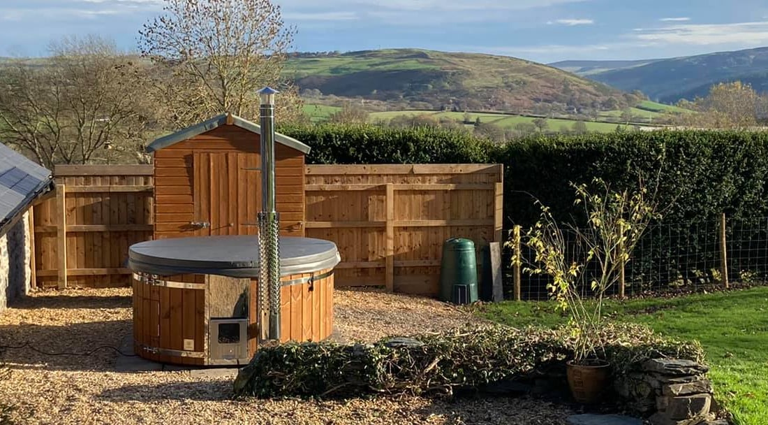 Outdoor view of a wooden hot tub with stainless steel chimney, set on gravel with garden shed and hills in background