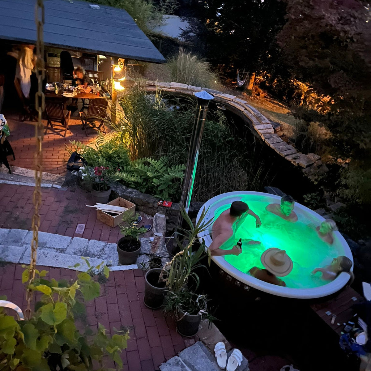 Evening view of a wood fired hot tub glowing green with four people relaxing in a garden patio setting with plants and seating area nearby