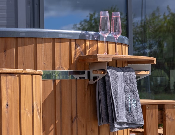 Gros plan sur le côté en bois de bain nordique avec plateau pliable contenant deux verres et une serviette suspendue gris foncé avec logo brodé