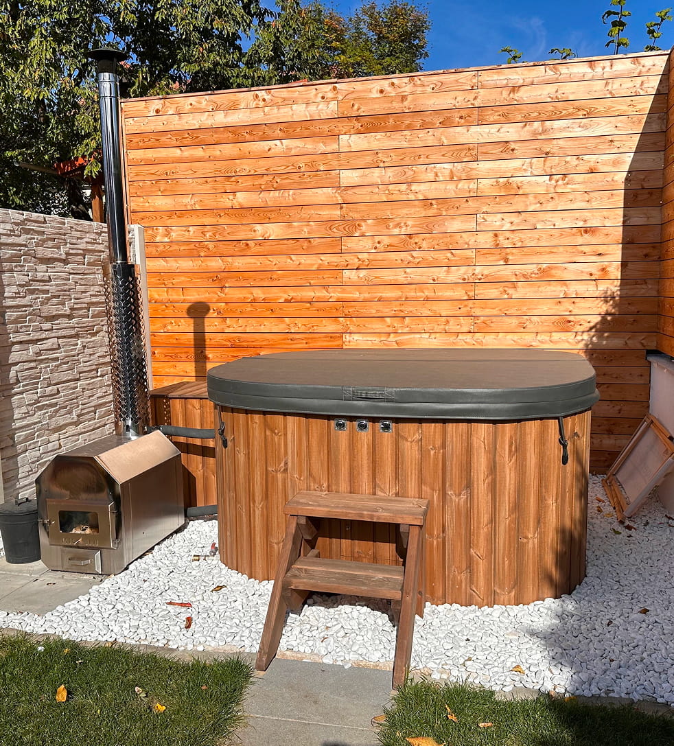 Square hot tub with a cover and standard step in a garden setting with a wooden fence and trees.